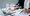 A close up of a woman's hands. One hand is on a laptop keyboard the other holds a pen and is checking notes on a clip board. Over the image are transparent symbols and the letters AI