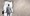 A businessman in a grey suit holding a very large sledge hammer is standing in front of a large hole he has smashed through a concrete wall
