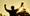 A man with the sun setting behind him is holding a medal in his hand and with his arms raised in victory