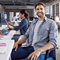 A man sitting at his desk and looking at us with a smile on his face. In the background is a work colleague and a large office space
