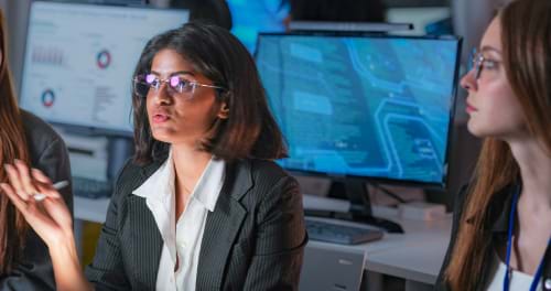 Business women sitting with their backs to their computer screens and looking across the room and talking with people off screen.